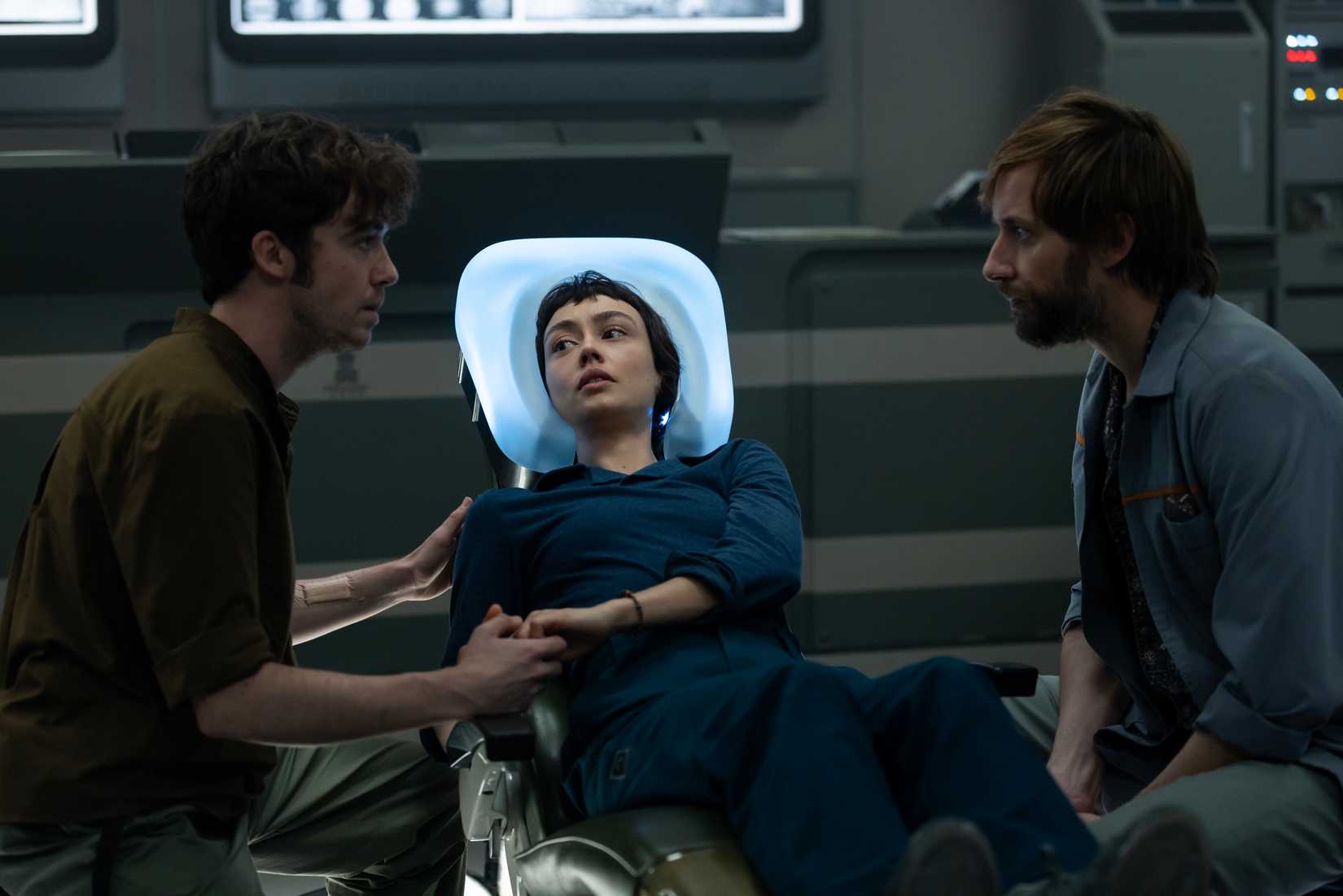 Hermit (Alex Lawther) and Arthur (David Rysdahl) sit close to Wendy (Sydney Chandler) as she lies on a medical bed looking worried and holding Hermit's hand in Alien: Earth