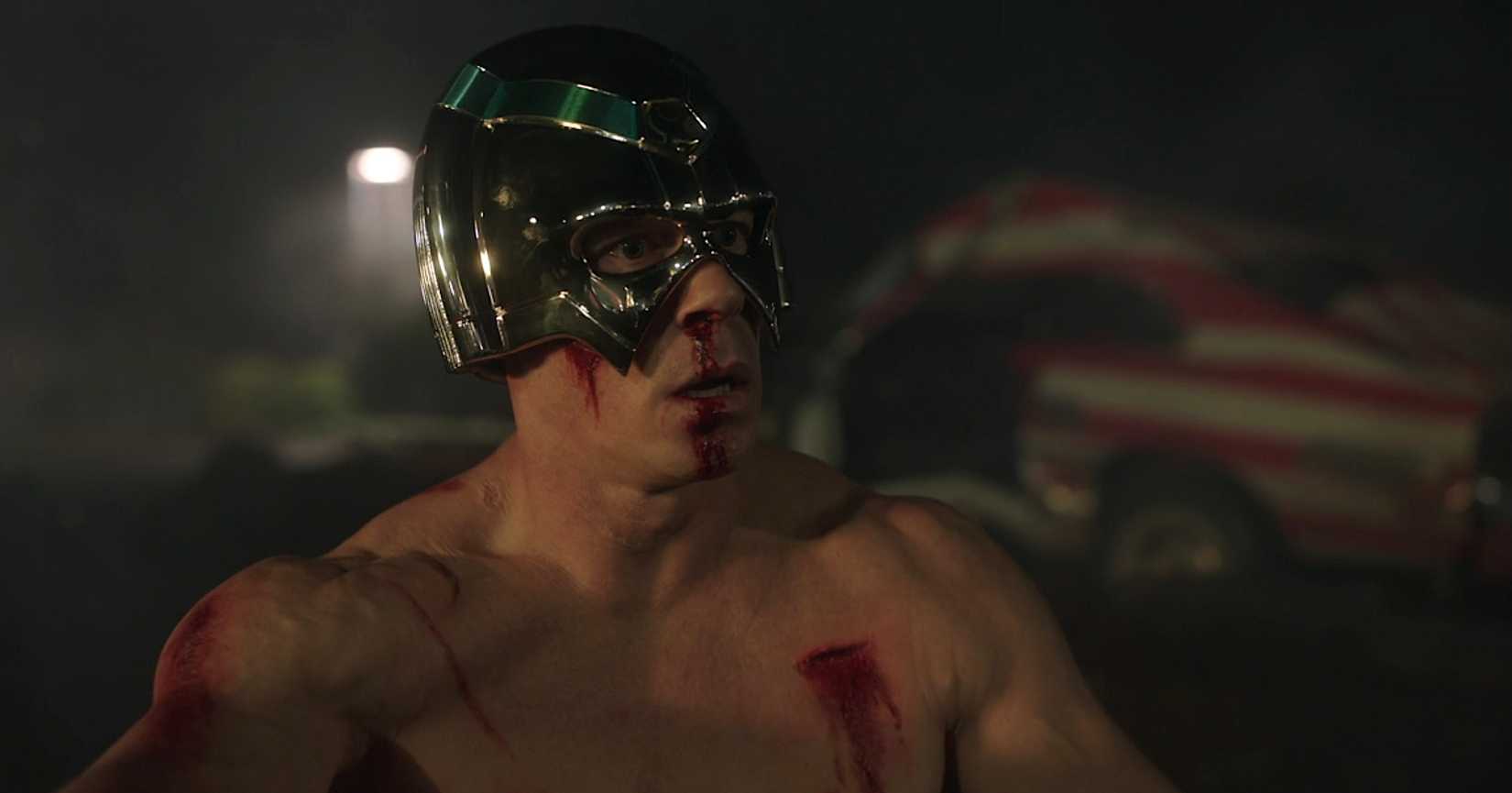 Peacemaker (John Cena), shirtless, stunned, and covered in bleeding wounds, sits in a parking lot at night in season 1, episode 1