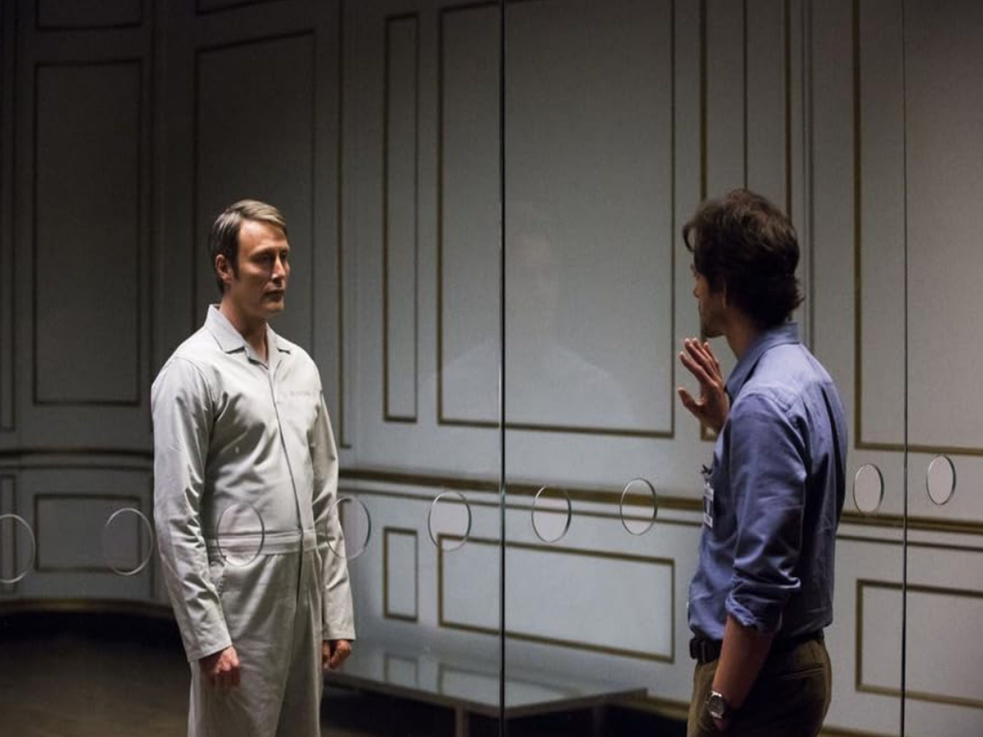 Mads Mikkelsen as Hannibal Lecter stands in front of a glass barrier. He wears a white prison outfit. On the opposite side of him is Hugh Dancy as Will Graham, wearinga blue shirt and trousers. Will has a hand pressed to the glass screen. Image from NBC's Hannibal by Bryan Fuller.