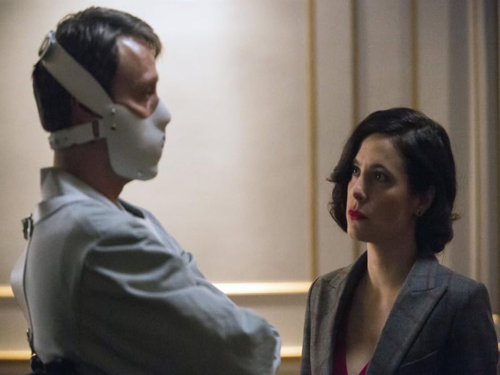 Mads Mikkelsen as Hannibal Lecter. He is dressed in a white prisoner outfit with a white muzzle over his face. Caroline Dhavernas as Alana Bloom watches him, wearing a grey pinsuit. Image from NBC Hannibal's by Bryan Fuller.