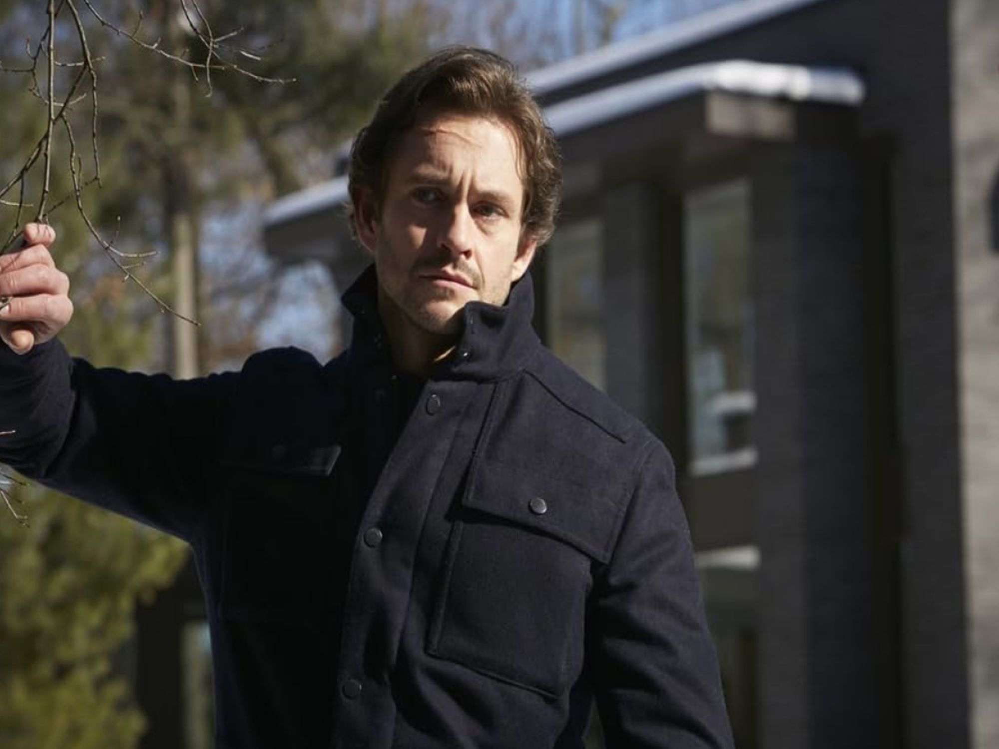 Hugh Dancy as Will Graham. He wears a dark coat and stands in front of a slate-gray building. From NBC's Hannibal.