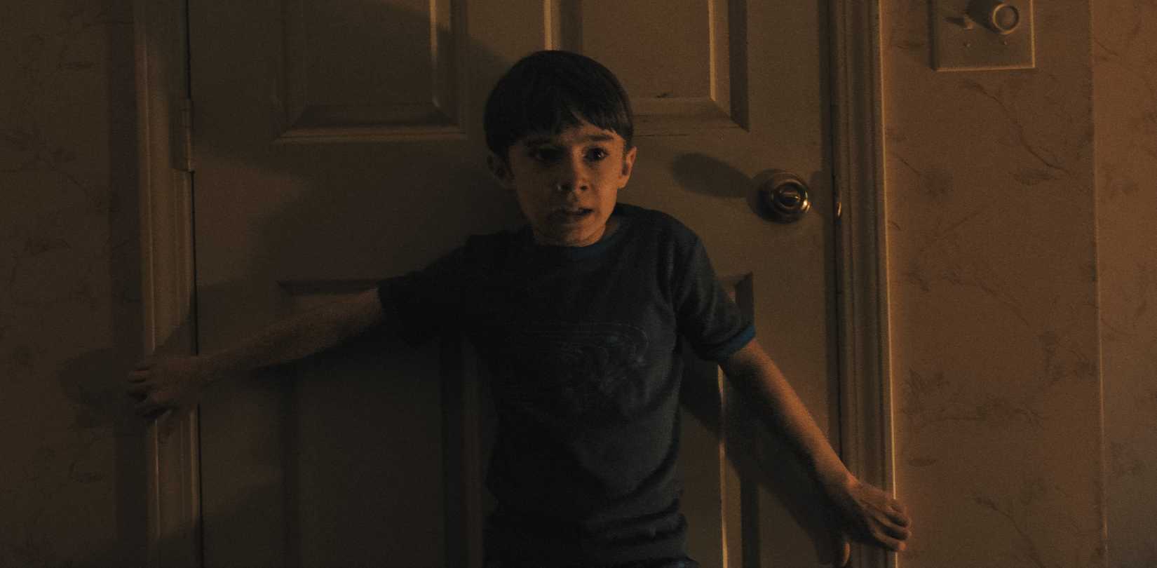 A young terrified boy stands with his back pressed to a closed door and his arms outstretched to block it in Weapons