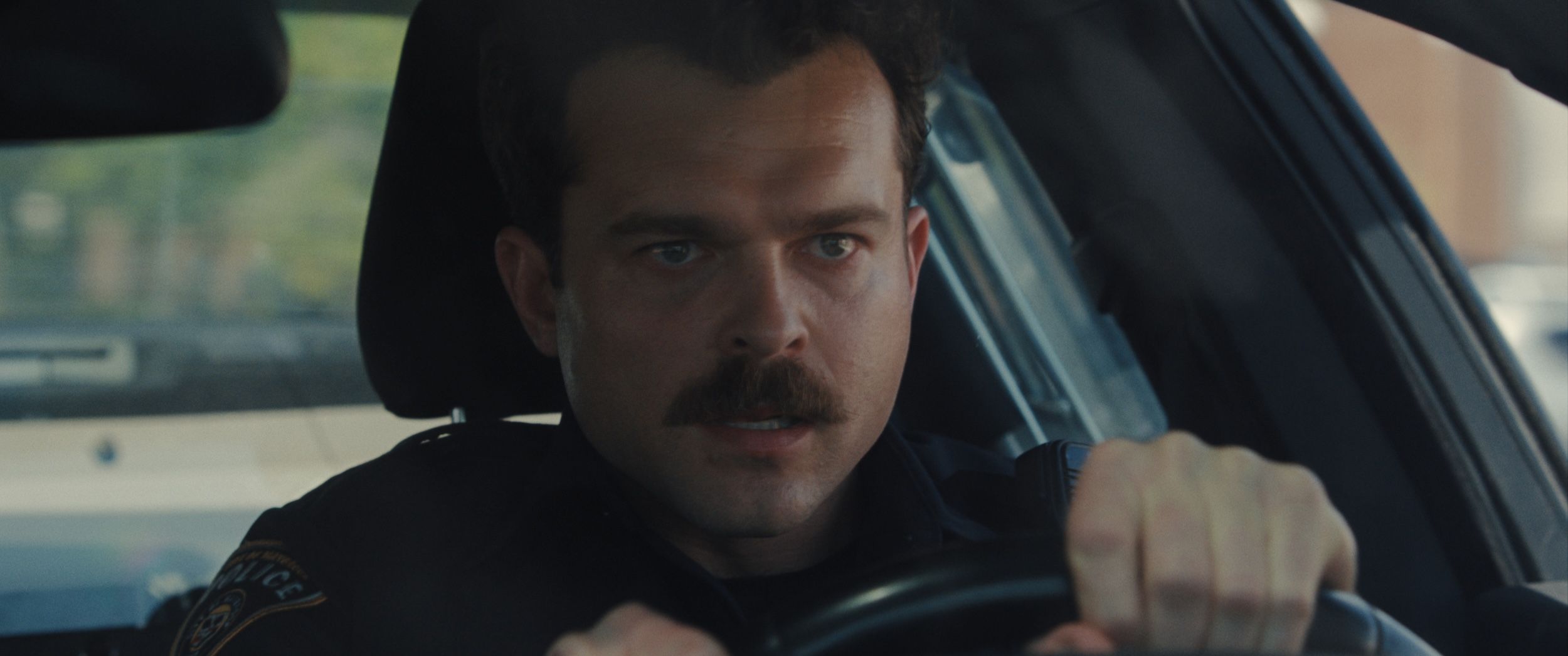 Alden Ehrenreich with mustache driving a police car in Weapons