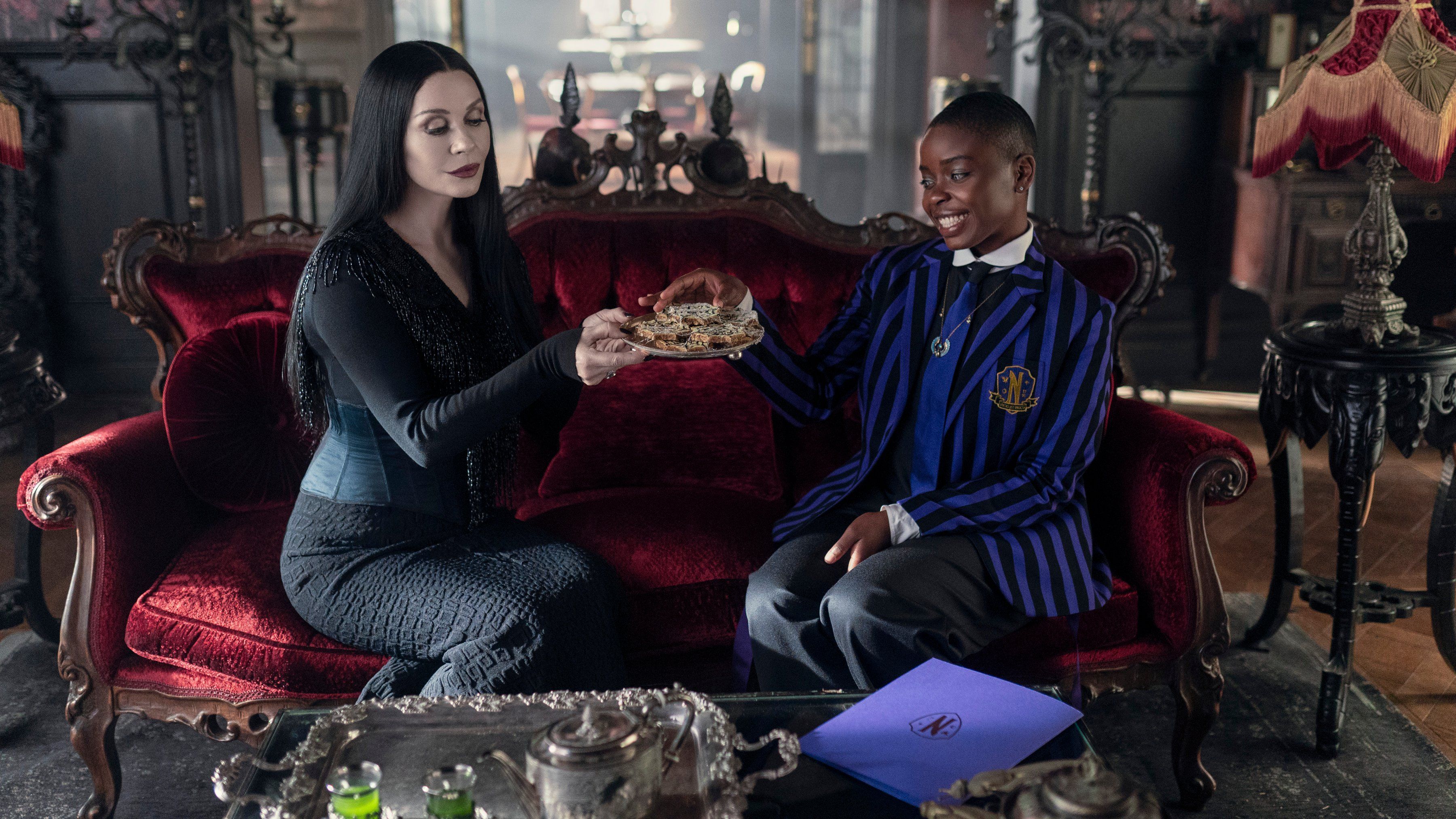 Catherine Zeta-Jones as Morticia Addams holds out a tray of cookies while sitting on a couch with Joy Sunday as Bianca Barclay in Wednesday