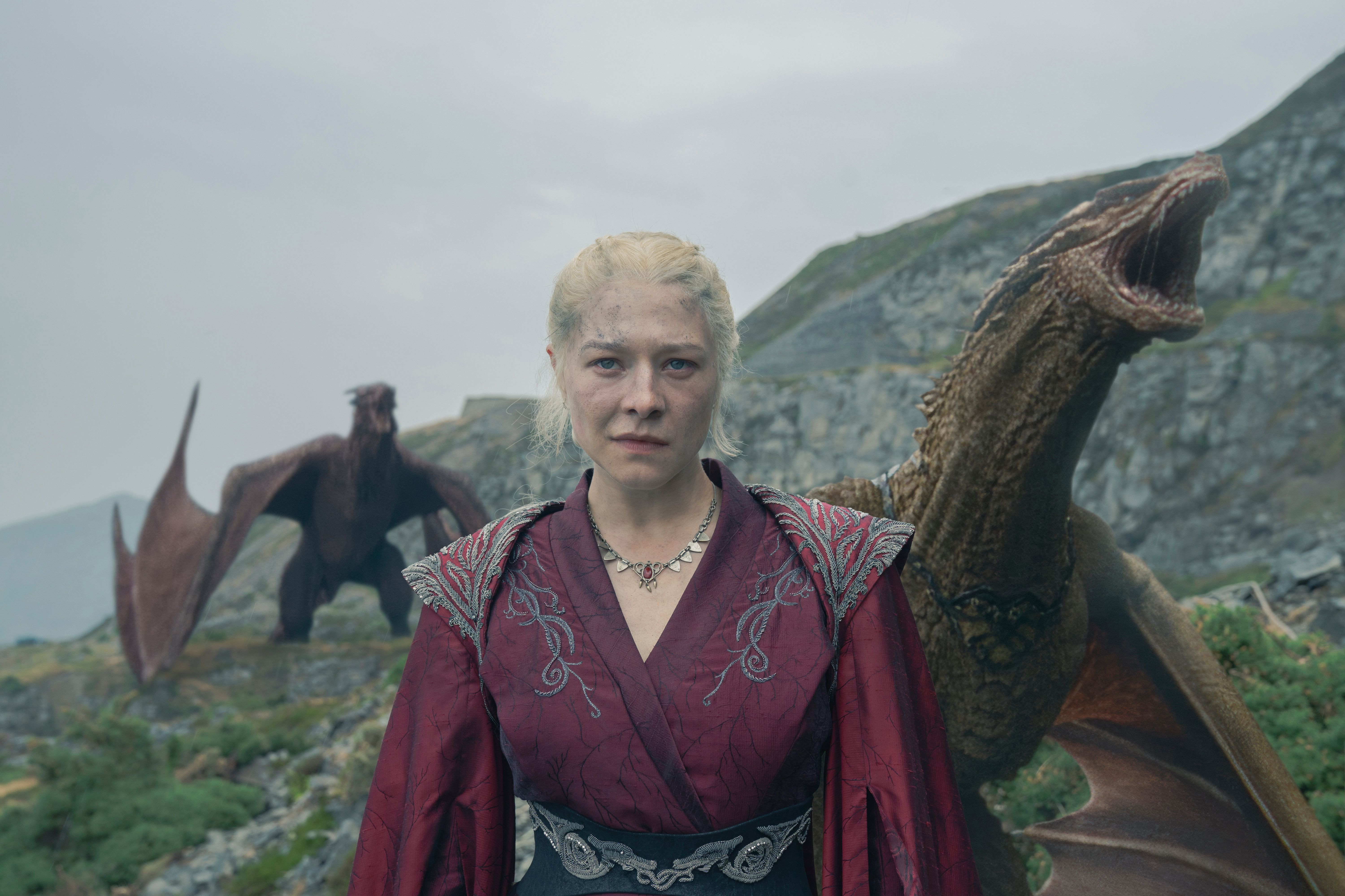 Rhaenyra se para frente a algunos acantilados rocosos mientras dos dragones rugen detrás de ella